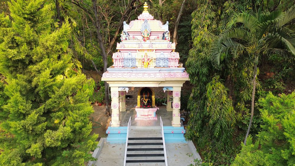 A rose-and-white Ganesha shrine framed by cypress and rain trees (Pink.lotus.deva, Wikimedia Commons · CC0).