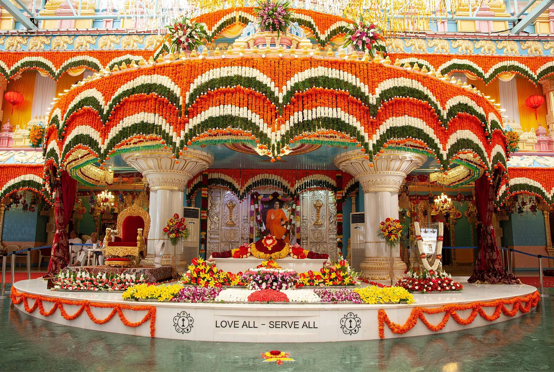 Inside Sai Kulwant Hall — the Mahasamadhi beneath arches of marigold, LOVE ALL — SERVE ALL on the dais
